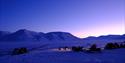 Persons on snowmobiles heading out on a trip with a mountainous landscape covered in blue light in the background