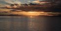 A dark fjord and dark skies with a setting sun and mountains in silhouette in the background