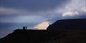 En person som står ved en varde på toppen av et fjell, i silhuett mot en fjord og et fjell i solskinn i bakgrunnen