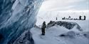 A person stands under a wall of ice looking up at the ice wall. In the background, several people and snowmobiles stand in a snowy landscape.