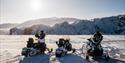 Three snowmobiles stand on the Arctic Ocean with a glacier in the background. Two people stand by their snowmobiles. The sun hangs over the mountains
