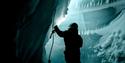 As a silouette, a person is standing inside a ice cave. Around the person, it's walls of blue ice, partly covered in snow.