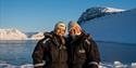 Two people smiling in front of an Arctic fjord landscape.