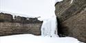 The picture is taken from below, looking up a mountain wall. The ground is covered with snow, and down from the mountain wall there is a frozen waterf