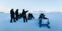 flere personer og snøscootere. snøscooterne står parkert og personene ser utover et snødekt landskap