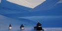 Three snow mobiles are driving in the winter landscape with big mountains in the background,