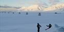 A small person walking up a small hill and another small person sliding down the snowy hill. With historic buildings and snow-covered mountains in the