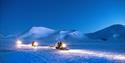 Tre snøscootere kjører på snøen med fjell i bakgrunnen og mørkeblå nattehimmel
