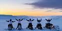 A group on snowmobiles raising their arms towards the sky at sunset.