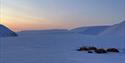 En teltleir i mørke snødekte omgivelser i forgrunnen, med fjerne fjell og lys fra en solnedgang på himmelen i bakgrunnen