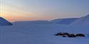 En teltleir i mørke snødekte omgivelser i forgrunnen, med fjerne fjell og lys fra en solnedgang på himmelen i bakgrunnen