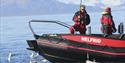 Two persons on board a RIB boat where one is holding a fishing pole with a fish on the hook floating on the water's surface in the foreground, surroun