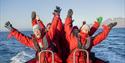 A group of guests sitting in a RIB boat at speed cheering with their arms outstretched