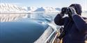 A guest on a boat in the foreground scouting out across a calm blue fjord and mountains with binoculars