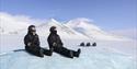 Two persons in snowmobile equipment sitting on an ice formation to relax in the foreground, with snowmobiles parked in a snowy landscape in the backgr