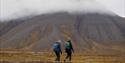 To gjester på fottur på tundraen med fjell og skyer i bakgrunnen