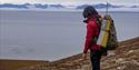 A guide with backpack and rifle on their back, looking out across a fjord in the background