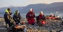 Two guides and two guests sitting on a beach talking with each other, and a RIB boat anchored by the shore of the fjord in the background