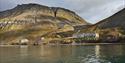 Forlatte bygninger i det tidligere gruvesamfunnet Grumant langs bredden av Isfjorden i forgrunnen, med høye fjell og en skyet himmel i bakgrunnen