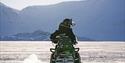 A guest wearing snowmobile equipment sitting on a snowmobile in a snow-covered landscape while looking behind themselves