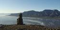 A cairn on top of a mountain with a fjord, a mountain and a clear blue sky in the background