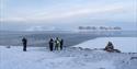 A guide and three guests in snowmobile equipment standing together in a snow-covered hilltop scouting out across a fjord with mountains in the backgro