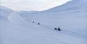 Three guests on snowmobiles in the background following a guide on a snowmobile in the foreground through a snow-covered landscape