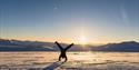 A person wearing snowmobile equipment doing a handstand in a snowy landscape with a clear blue sky and sunshine in the background