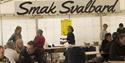 People eating lunch at tables in a festival tent, with a large "Taste Svalbard" logo and students making food on tables in the background