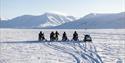 A guide standing in front of four snowmobiles with guests telling them about the snowy surroundings they're in, with a mountain range and a long snow-