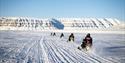 Four snowmobiles with guests following a guide on a snowmobile in the background through a snow-covered landscape towards a range of mountains in the