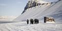 A group of guests in snowmobile equipment walking towards a building in snowy surroundings next to Villa Fredheim along Tempelfjorden, with mountains