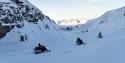 To gjester og en guide på snøscootere som kjører gjennom en mørk og trang snødekt elvedal med en blå himmel og fjell i solskinn fjernt i bakgrunnen