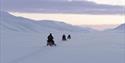 To gjester på hver sine snøscootere som følger en guide på snøscootertur gjennom et vidt åpent snødekt landskap