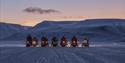 A group of guests on snowmobiles parked next to each other in a snow-covered valley in twilight conditions