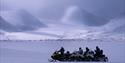 A guide and guests on snowmobiles in a landscape with mountains in the background