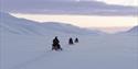 To gjester på hver sine snøscootere som følger en guide på snøscootertur gjennom et vidt åpent snødekt landskap