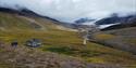 A cabin in a green mountainous landscape in the foreground, with a river and a glacier in between mountains in the background