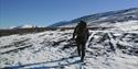 A guide with a rifle and backpack hiking through a mountainous landscape