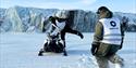 A snowmobile infront of a wall of blue ice in the background. One person is standing on one foot on the snowmobile. Another person is walking towards