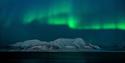 A darkened river and mountain formation are illuminated by the northern lights dancing across the sky.