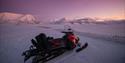 A red and black Lynx snowmobile is parked for a photo with the mountain formations and the purple sky in the background.