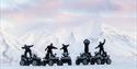 5 people standing on 5 ATVs. Mountains in the background.