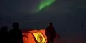 People standing outside a lighted tent in the dark at Svalbard, watching the northern lights.