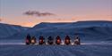 Seven snowmobiles have come to a stop side by side to observe the delicate mountain formations against an orange sky.