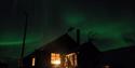 Cabin with light through the windows under the Northern Lights sky.