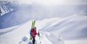 A person stands on a mountain ridge with skis in their backpack, with a sunlit mountain landscape in the background