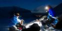 Two people with headlamps and a dog in the foreground, with mountains and Longyearbyen in the background.