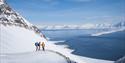 Three people stand on a mountain ledge, looking out over the fjord with skis on their feet.