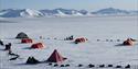 En teltleir med sledehunder, parkerte hundesleder og personer i et stort åpent snødekt landskap med fjell fjernt i bakgrunnen
A tent camp with sled do